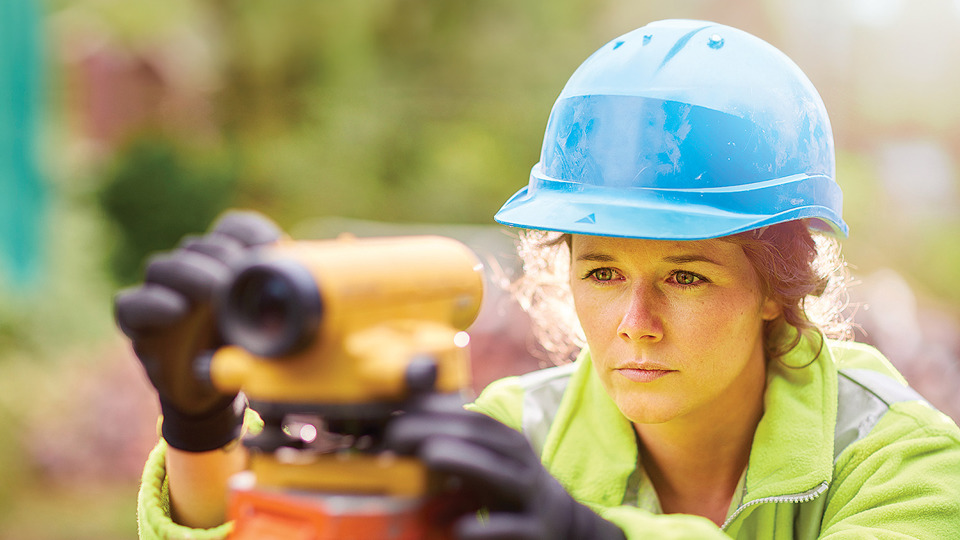 Woman wearing hard hat and high-vis jacket looking through levelling telescope