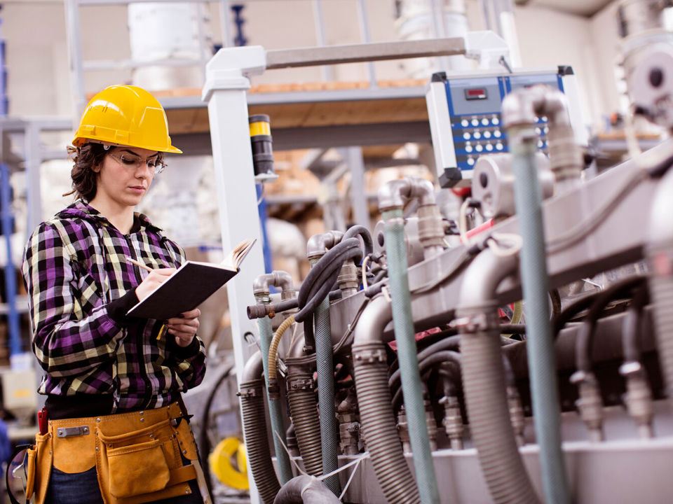 Woman in hard hat standing next to factory machine and making notes on clipboard